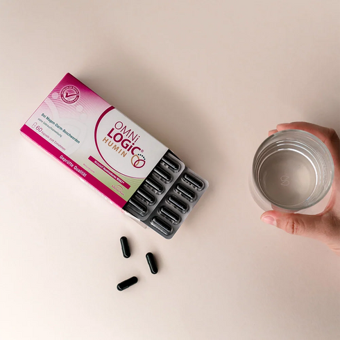 Eine offene Packung OMNi-LOGiC HUMIN mit losen Kapseln und einem Glas Wasser